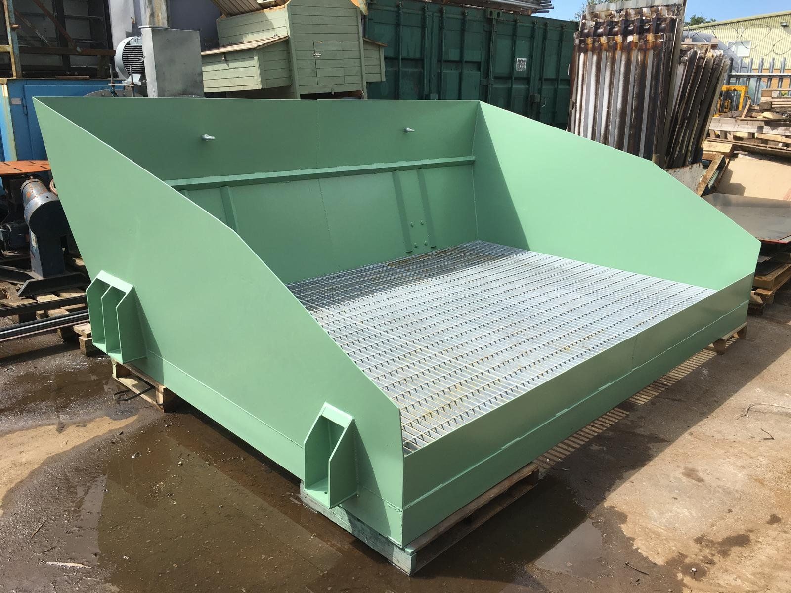 Green industrial machine with metal grating on the bottom, situated outdoors on a wooden pallet, with various metal parts and equipment in the background.