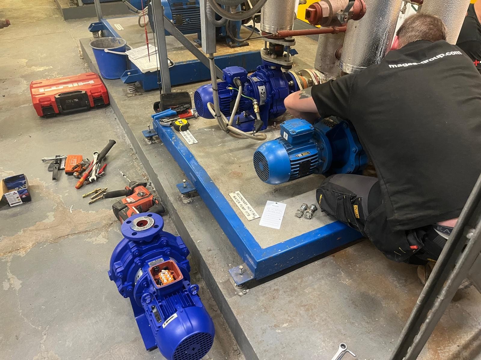 A technician working on a water pump system on a metal platform, with various tools and equipment around, on an industrial concrete floor.