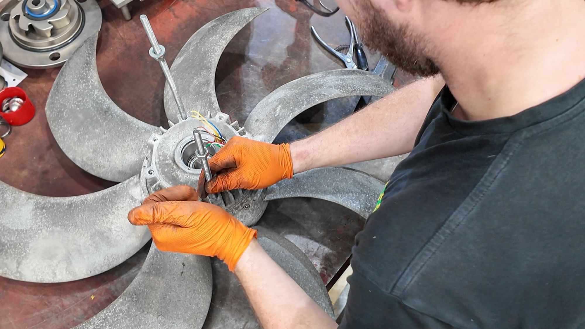 A person wearing orange gloves working on a metallic turbine blade with wires and mechanical parts, using a tool, on a workshop table.