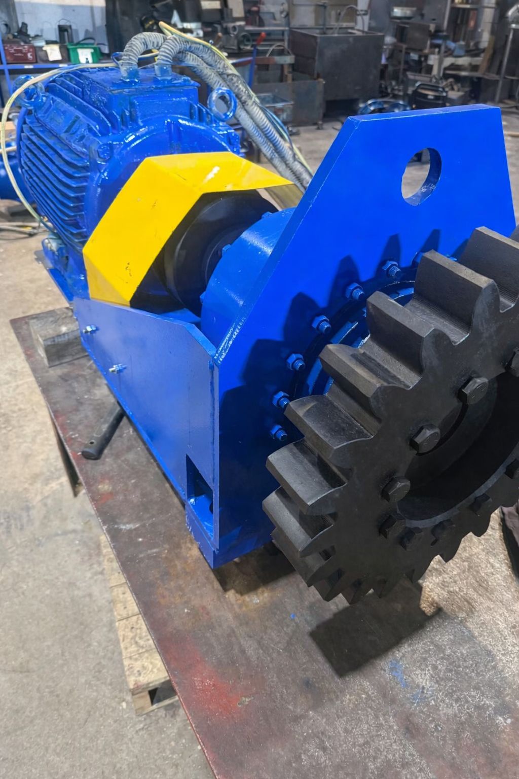 A large heavy-duty industrial machine with a blue and black color scheme, featuring a gear with teeth on one side, mounted on a workbench in a workshop.