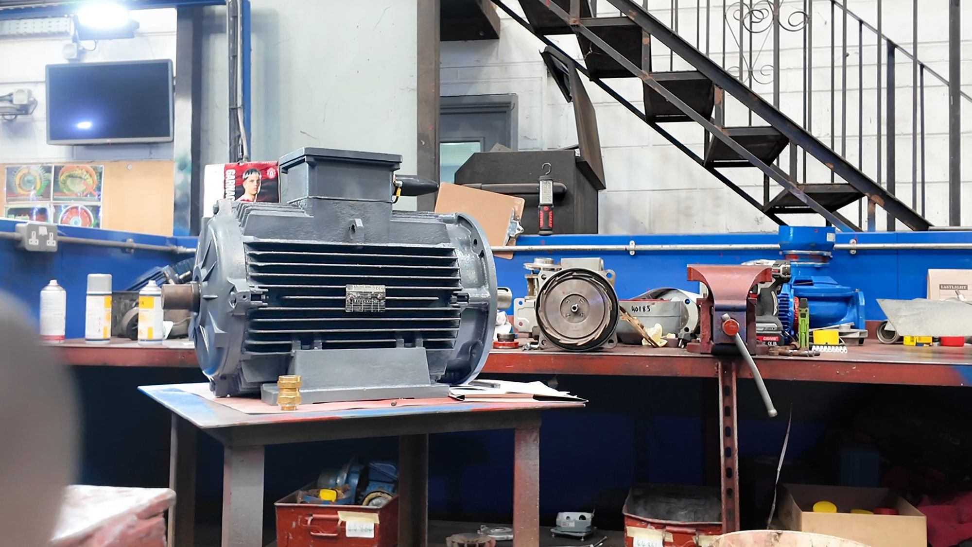 Workshop with machinery parts, tools, and supplies on a workbench, including a large motor or gearbox in the foreground, with various containers, a grinder, and equipment on the table and shelves behind.