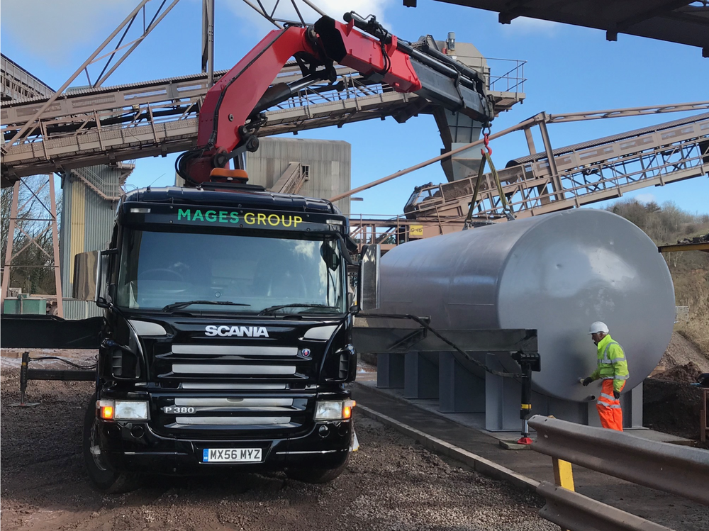 Construction site with a black Scania truck parked near a large cylindrical tank. A worker in a safety helmet and high-visibility orange pants is standing near the tank, and a red crane arm is lifting materials overhead. Industrial structures and conveyor belts are visible in the background against a blue sky.