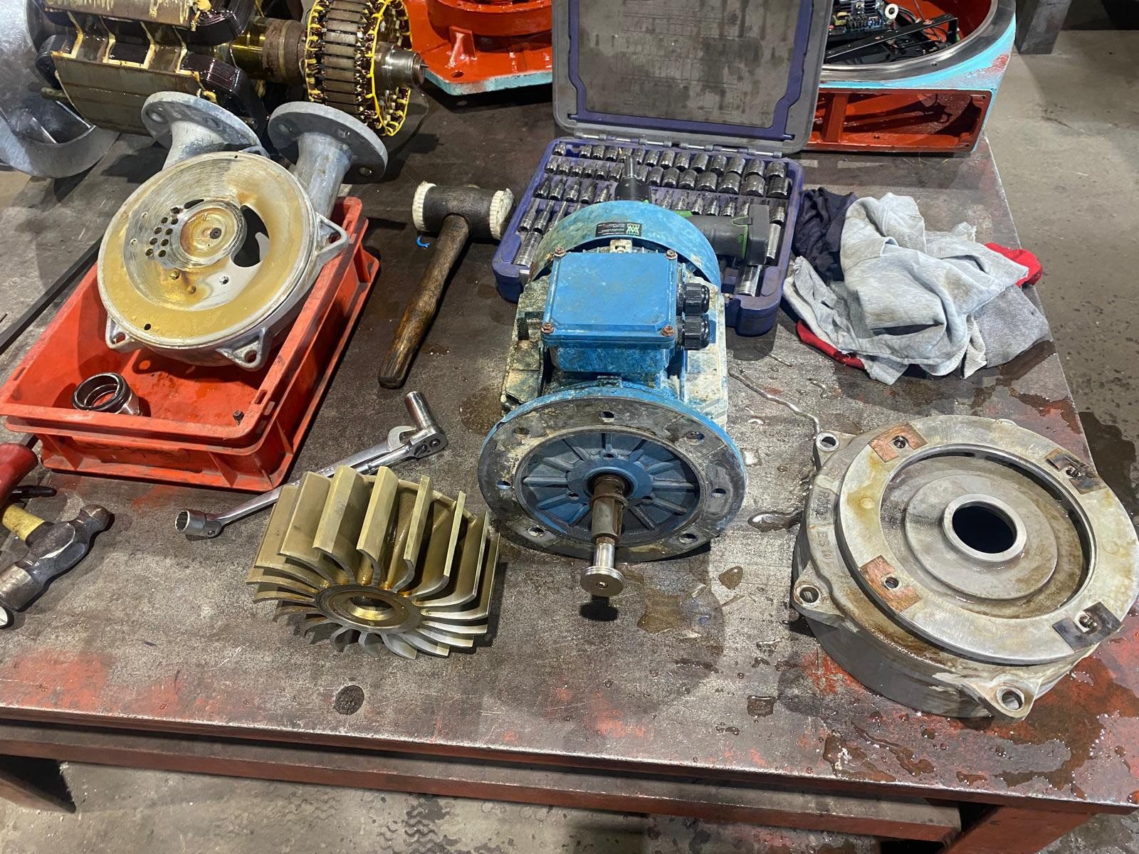 Disassembled automotive parts on a workbench, including gears, a blue electric motor or power tool, and tools scattered around.