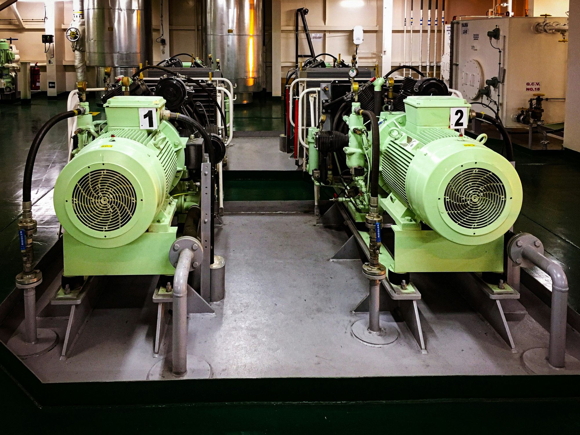 Two large, green industrial compressors labeled '1' and '2', connected with black hoses, situated in a factory or industrial setting with other machinery and equipment in the background.